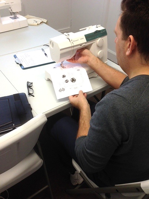 Student practising sewing machine control during a workshop, guiding fabric under the needle with other machines and materials on the table.