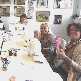 A group of 3 students at a sewing class sitting around a table. They are grinning and holding up zip; having learned how to insert them.