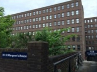 A large brick building with multiple rows of windows and a blue sign reading “St Margaret’s House” at the entrance, with bicycles and greenery along the walkway.