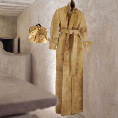 The image shows two pieces of clothing made from Reishi fabric, which was unveiled at Paris Fashion Week in 2023. The larger piece is a long coat with a belt and cuffs, and the smaller piece is a vest. Both garments are hanging against a textured, beige wall in a minimalist setting. The Reishi fabric has a unique, earthy appearance, highlighting its innovative use in fashion.