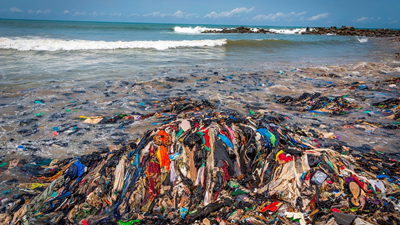 a tropical beach, with an azure sky and jade green water, a scene of paradise, but piled high with discarded waste clothing from the fashion industry as far as can be viewed.