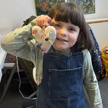a little girl is giving a cheeky grin and holding up a heart-shaped pin cushion full of colourful pins. She is wearing a denim pinafore-style apron that she & her mum made during the Family Sewing class. The Apron is upcycled from a pair of adult jeans.