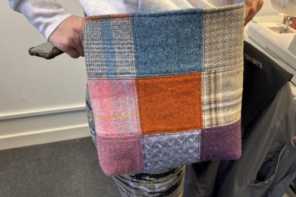 A handmade patchwork crossbody bag made from colourful tweed squares, held by a person standing beside a sewing machine in a studio.
