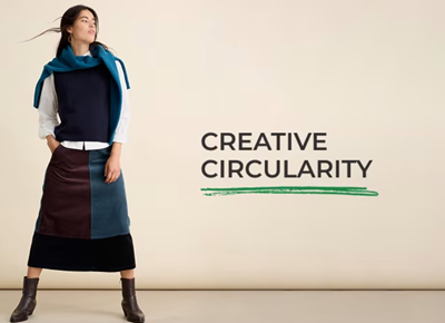 A woman is dressed in a white cotton shirt, and a blue skirt made from panels of different fabrics from upcycled garments. Across her shoulders is slung a cotton sweater, and she wears brown leather ankle boots