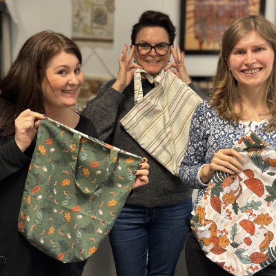 Beginner sewing students hold up their tote bags proudly. They are smiling and laughing, having enjoyed their sewing class