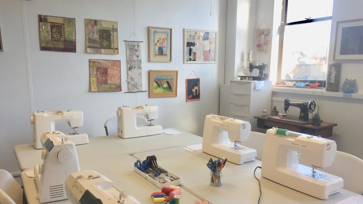 Light-filled sewing studio at Artisan Stitch in Edinburgh with sewing machines arranged for teaching, work tables prepared for class, and colourful textile art displayed on the walls.